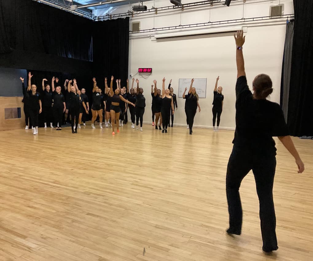 A group of students dancing in a studio, all with one arm up